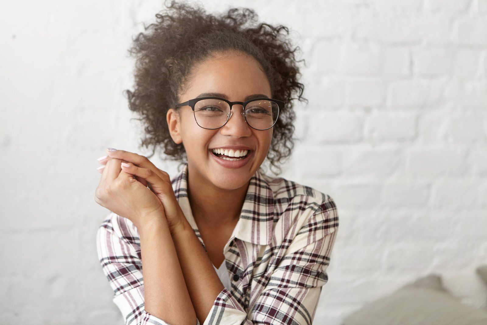 Youth and happiness concept. Attractive African American student girl with curly hairstyle spending nice time indoors, sitting against white brick wall, looking and smiling happily at camera